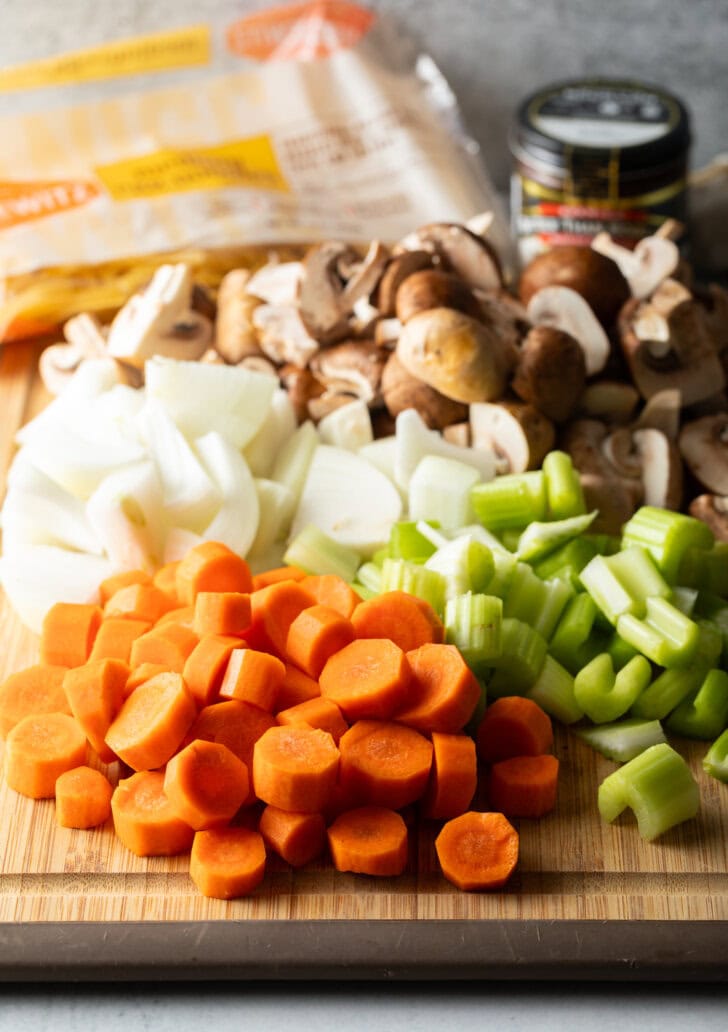 Soup ingredients, chopped in individual piles on a wood cutting board are a pile of carrots, onions, celery, and mushrooms.
