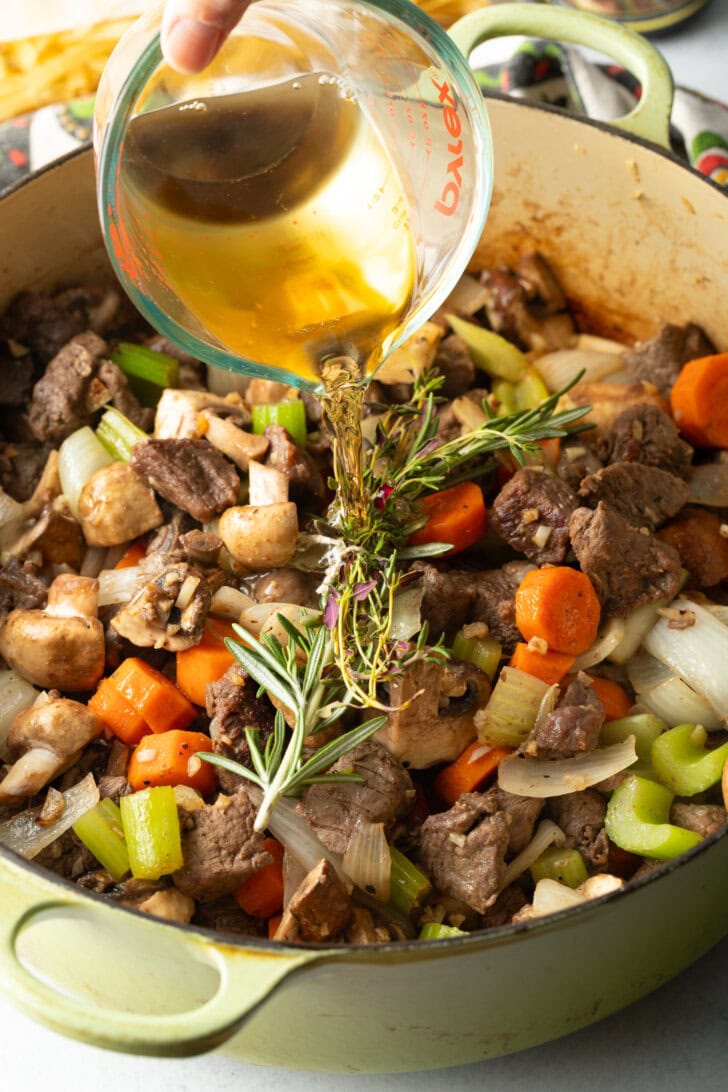Searing beef chunks and veggies in a large cooking pot. Adding sprigs. Pouring in broth