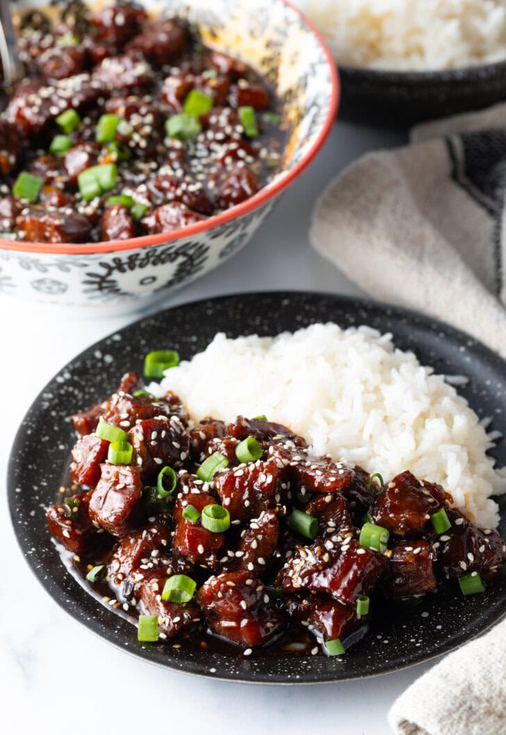 Bowl of beef teriyaki garnished with sliced green scallions, with white rice side-by-side in the bowl.