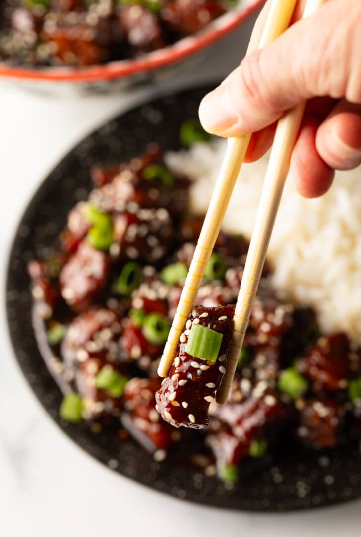 Bowl of beef teriyaki garnished with sliced green scallions, with white rice side-by-side in the bowl. A hand with chopsticks is holding a bite to camera.