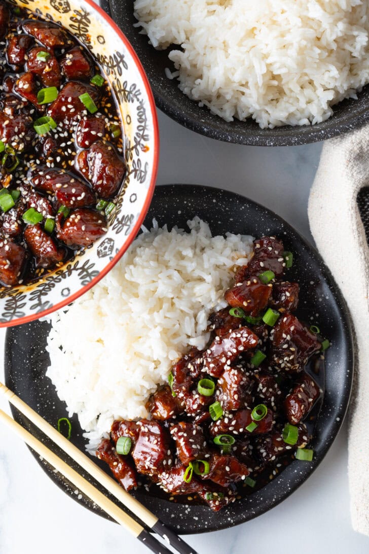 Bowl of beef teriyaki garnished with sliced green scallions, with white rice side-by-side in the bowl.