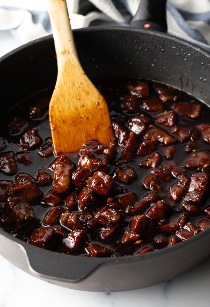 Deep black skillet with chopped beef in a brown sticky teriyaki sauce. A wooden spoon is in the skillet.