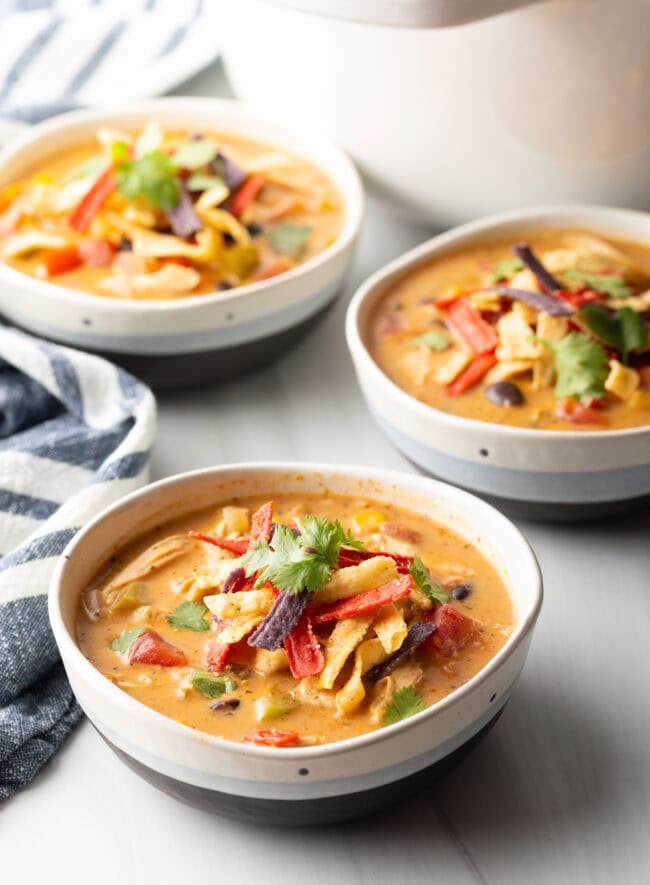 Three white and gray bowls filled with chicken taco soup. Each bowl is garnished with cilantro and red tortilla strips.