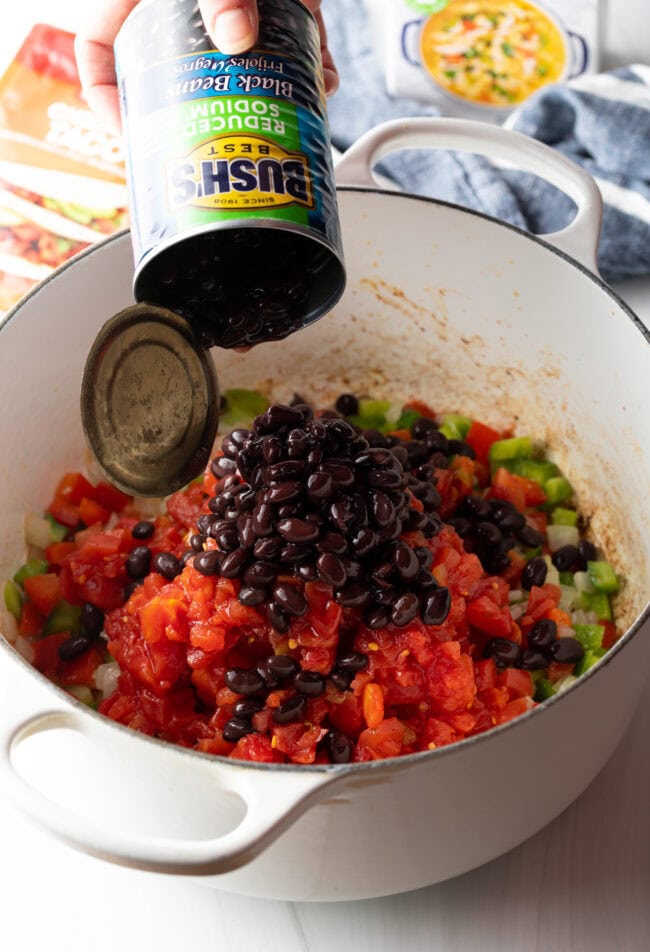 Hand adding canned black beans to pot with chopped veggies.