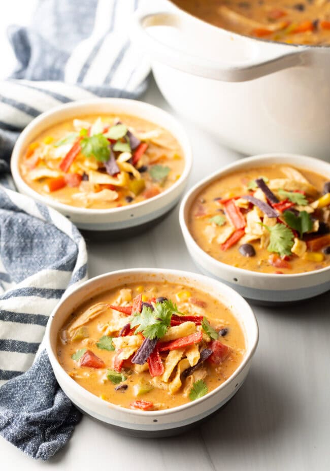 Three white and gray bowls filled with chicken taco soup. Each bowl is garnished with cilantro and red tortilla strips.