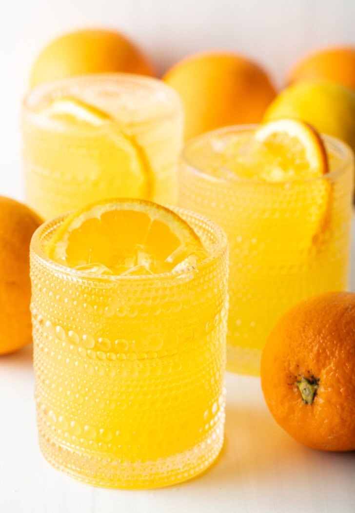 Two orange crush drinks in glasses, each garnished with a slice of orange. There are fresh oranges and lemons around the glasses.