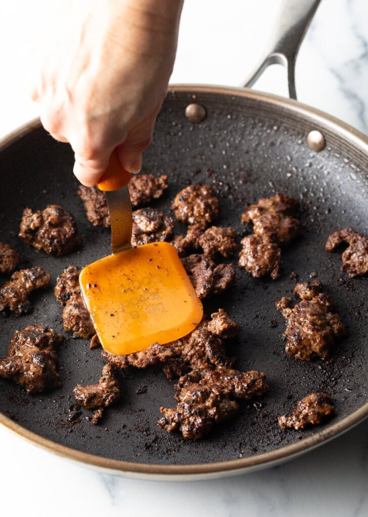 Hand with spatula cooking mini burgers in a skillet.