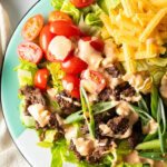 Big mac burger salad with shredded lettuce, ground beef, halved grape tomatoes, shredded cheese, pickles, and sliced green onion, all topped with a drizzle of Thousand island dressing.