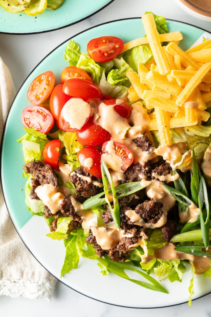 Big mac burger salad with shredded lettuce, ground beef, halved grape tomatoes, shredded cheese, pickles, and sliced green onion, all topped with a drizzle of Thousand island dressing.