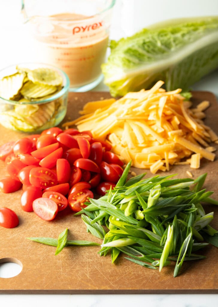 Recipe ingredients on a cutting board: Piles of shredded cheese, halved tomatoes, sliced green onions, and a small glass of pickle slices.