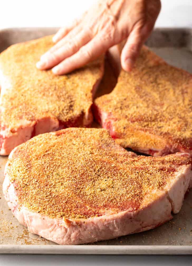Hands adding spice rub to 3 raw steaks.