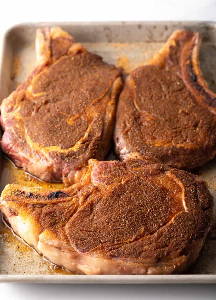 3 seasoned steaks with bones on a rimmed baking sheet.