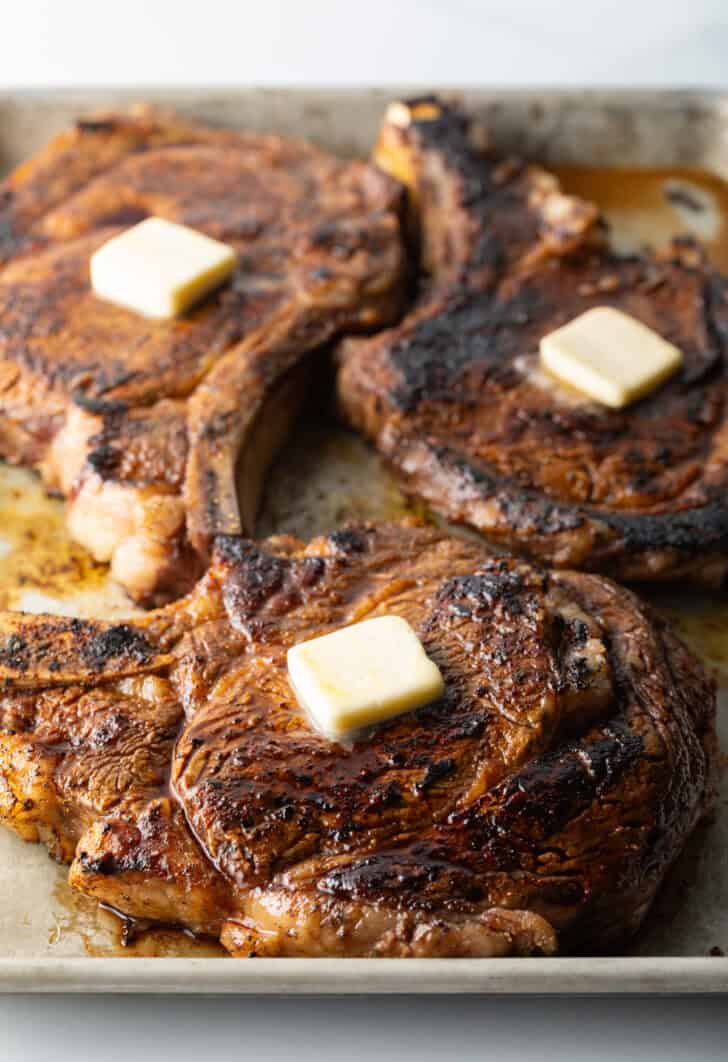 3 cooked steaks on a baking sheet, each with a pat of butter on top.