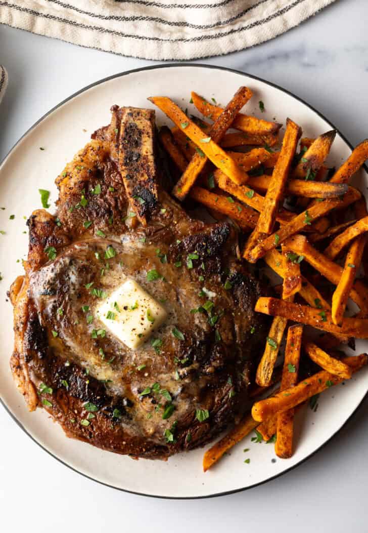 Large bone in ribeye steak on a plate with sweet potato fries.