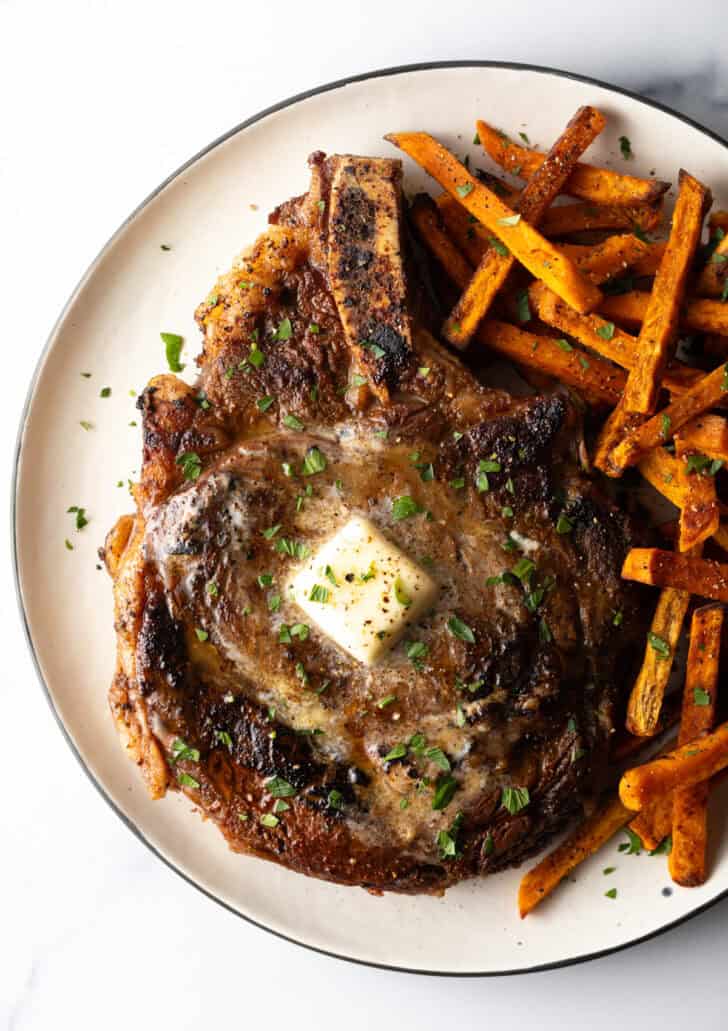 Large bone in ribeye steak on a plate with sweet potato fries.