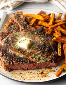 Large bone-in ribeye steak on a plate with sweet potato fries. The steak has been sliced to show the medium rare meat inside.