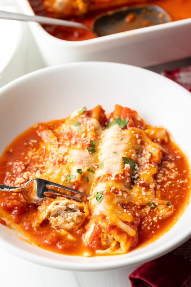 Two pieces of Manicotti Recipe with Meat in a white pasta bowl. A fork is taking a bit from one tube.
