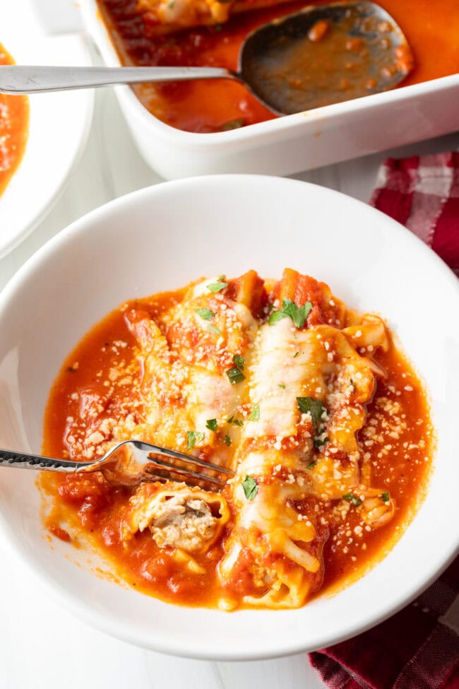 Two pieces of cheese manicotti with meat in a white pasta bowl. A fork is taking a bit from one tube.