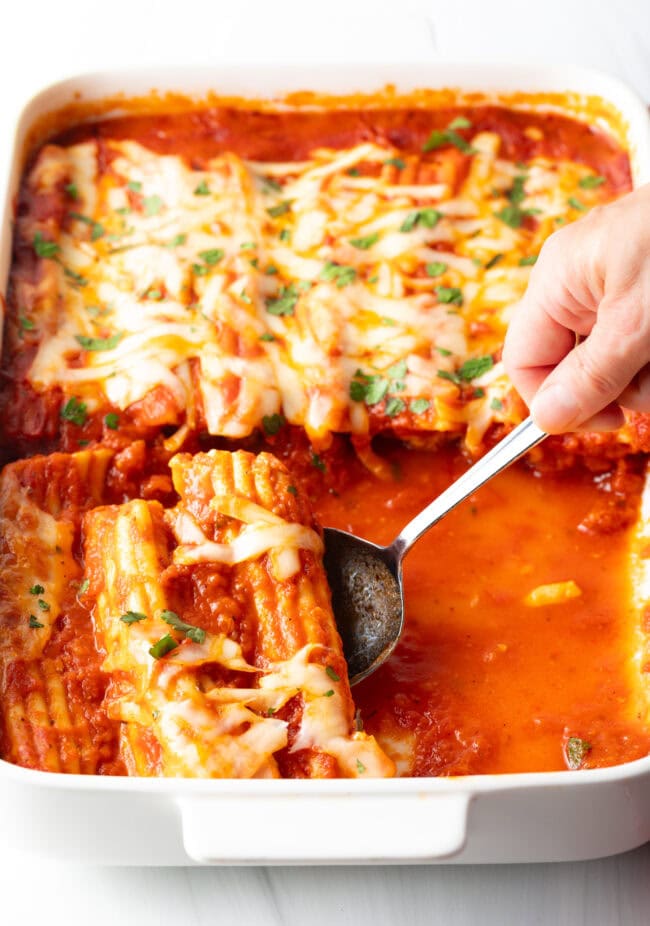 Hand with large metal spoon, scooping a serving of meat and cheese manicotti from the baking dish.