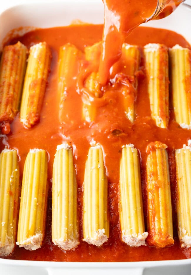 Pouring tomato sauce over tubes of stuffed pasta in a baking dish.