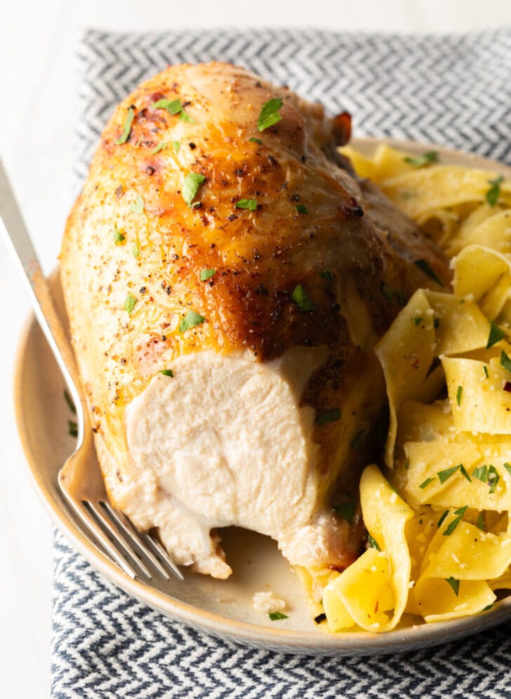 Bourbon brined chicken breast on a white plate with side of buttery noodles. A large bite has been removed from the chicken to show the moist interior.