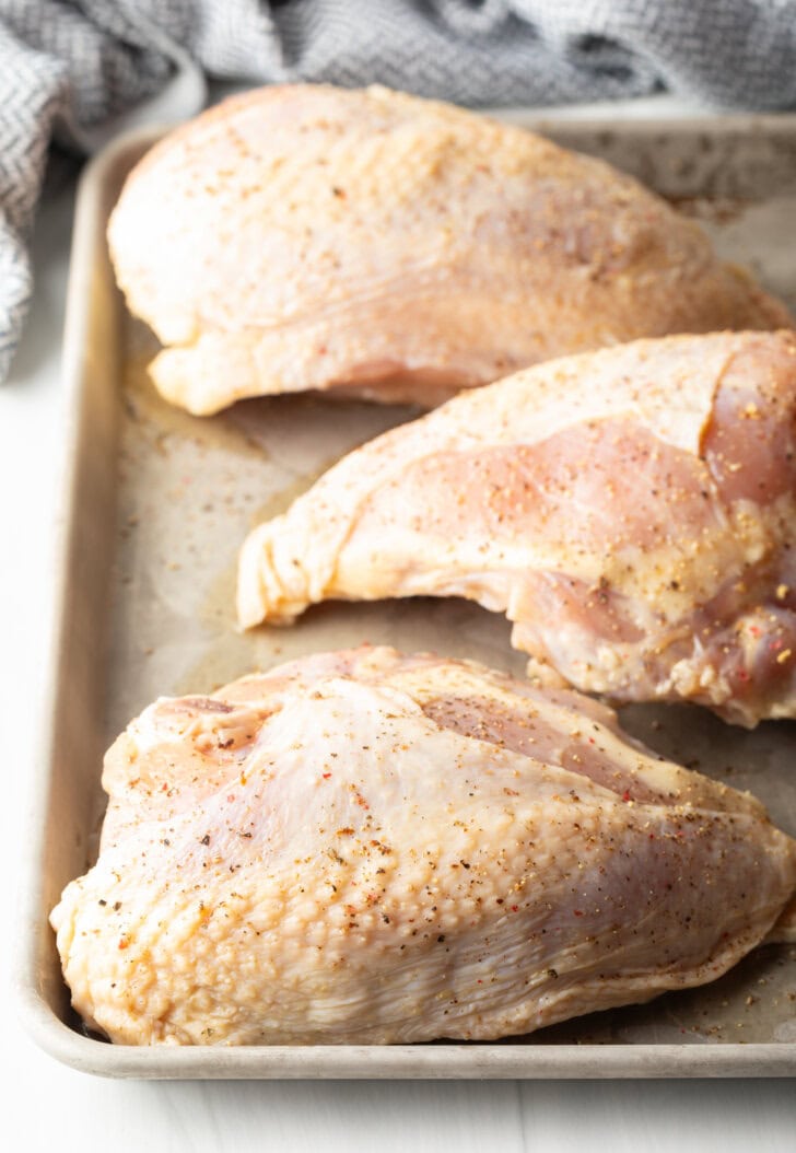 3 raw skin-on chicken breasts on a baking sheet.
