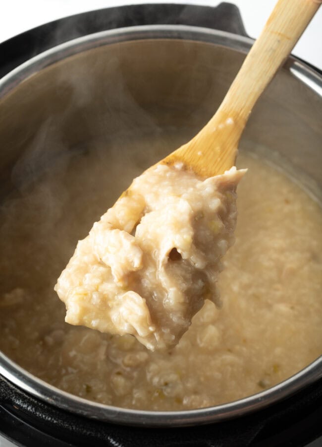 Wood spoon showing cooked chicken congee in instant pot to camera. Instant pot congee almost done cooking.