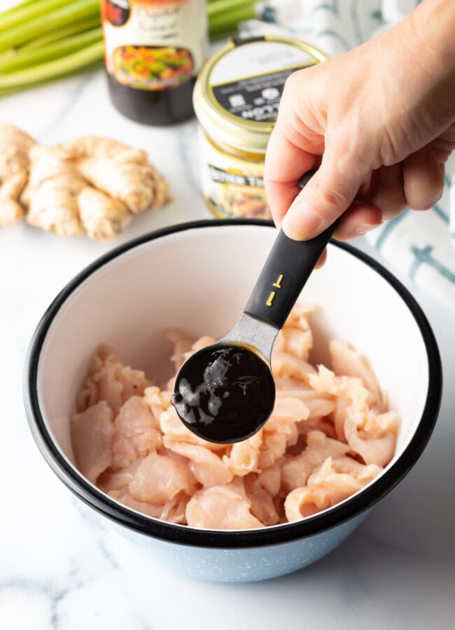 Hand with tablespoon filled with oyster sauce, adding to bowl with chopped raw chicken.