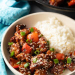 Top down view of white bowl, one half has white rice and the other half has a portion of manchurian chicken topped with sesame seeds and chopped green onion.