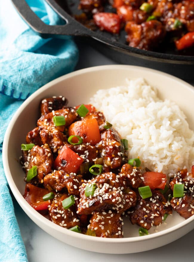 Top down view of white bowl, one half has white rice and the other half has a portion of manchurian chicken topped with sesame seeds and chopped green onion.
