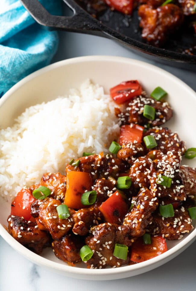 Top down view white bowl, one half has white rice and the other half has a portion of manchurian chicken topped with sesame seeds and chopped green onion.