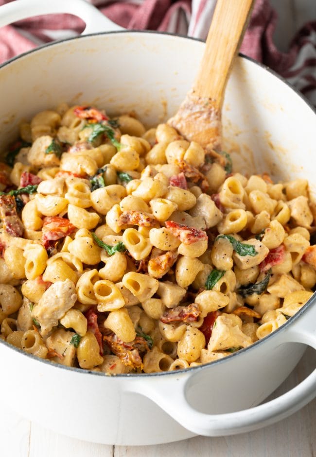 White pot of creamy macaroni noodles with chicken, sun dried tomatoes, and spinach.