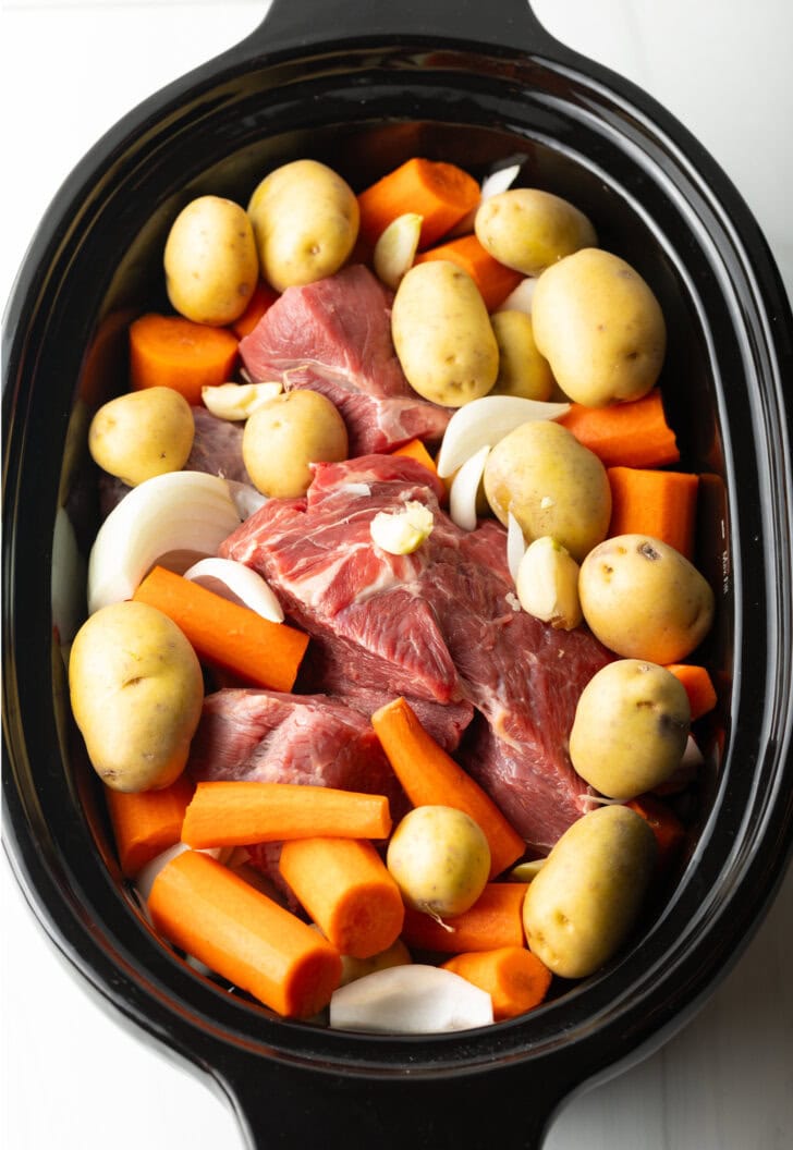 Top down in a slow cooker: Large raw beef roast is topped and surrounded by whole baby potatoes, chunks of carrots.