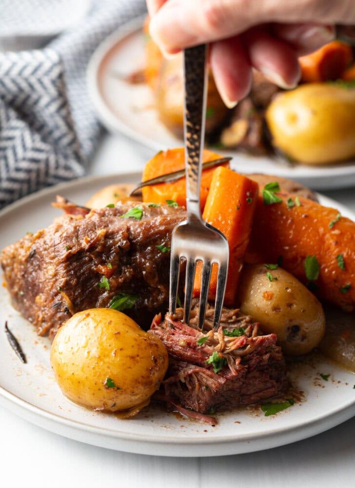 White plate with serving of beef pot roast, chunks of carrots, and whole baby potatoes. A fork is taking a bite of potato and chuck roast, and showing to camera.
