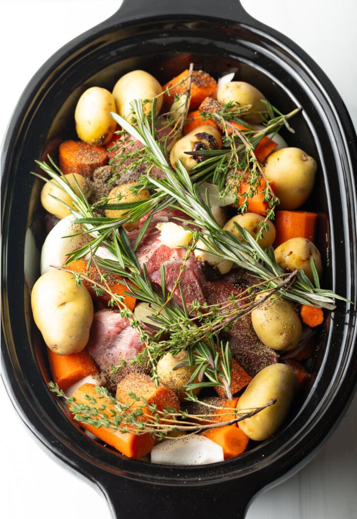 Top down in a slow cooker: Large raw beef roast is topped and surrounded by whole baby potatoes, chunks of carrots, and fresh thyme and rosemary sprigs.