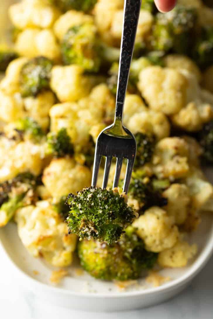 Fork holding a roasted piece of broccoli to camera, with a platter of roasted cauliflower and broccoli in the background.