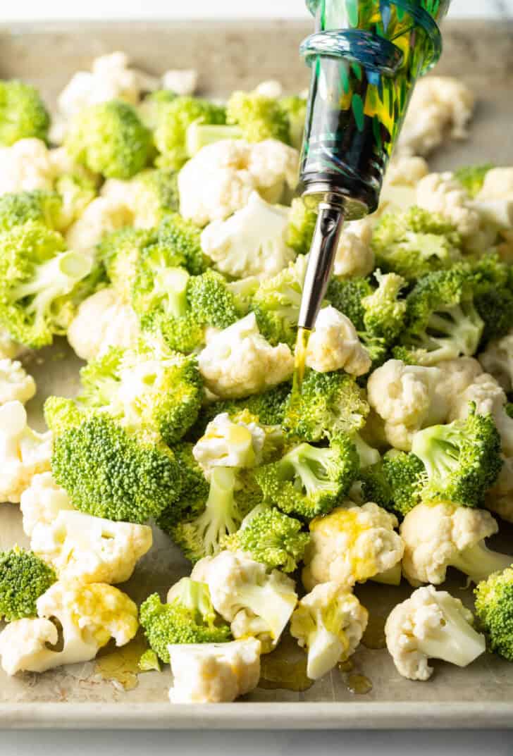 Adding oil onto raw broccoli and cauliflower florets on a baking sheet.