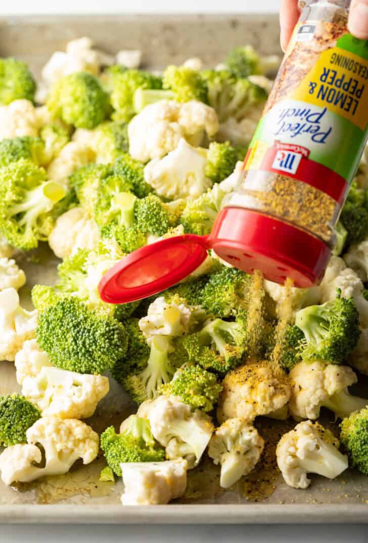 Sprinkling lemon pepper seasoning over broccoli and cauliflower florets on a baking sheet.