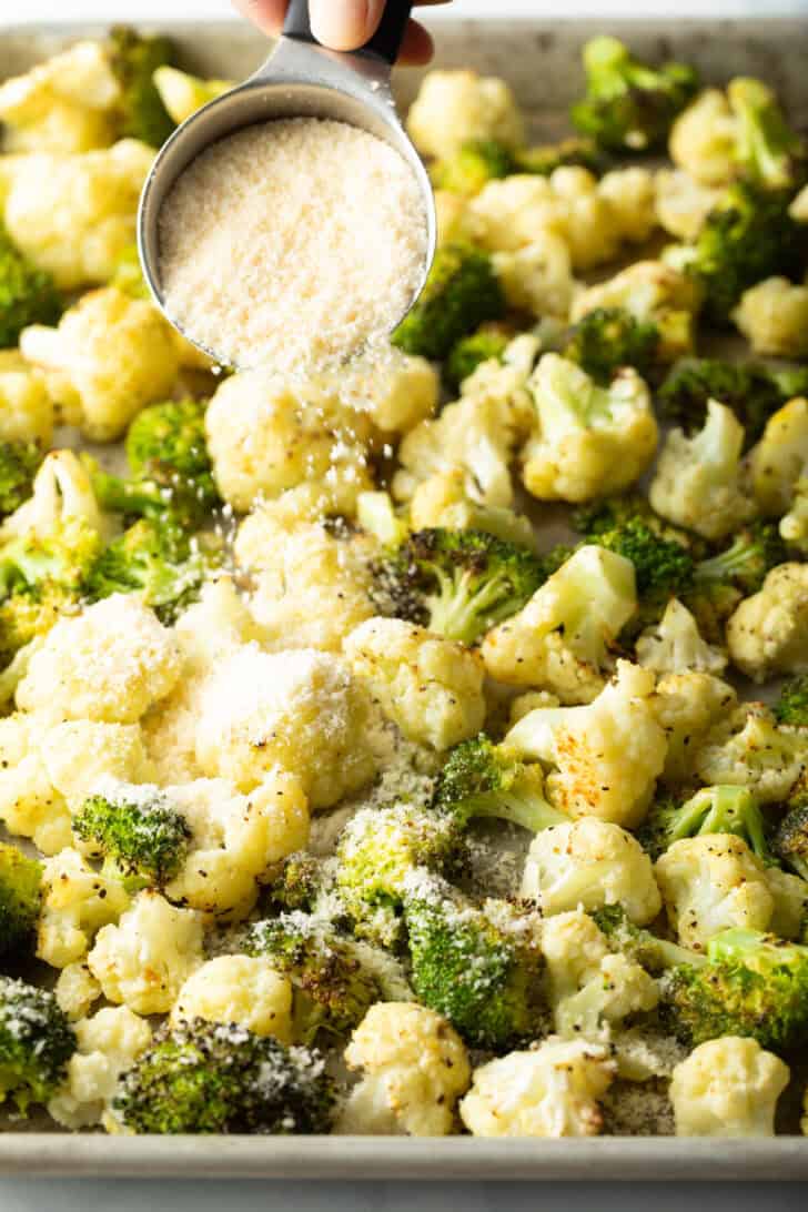 Sprinkling parmesan cheese over roasted pieces of broccoli and cauliflower on a baking sheet.
