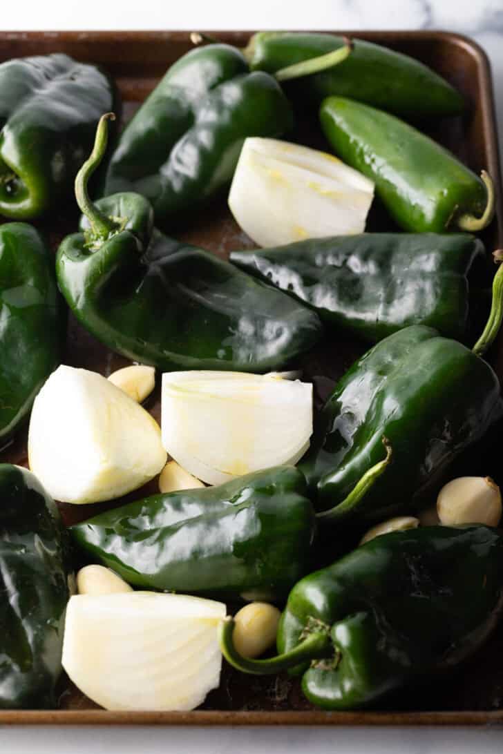 Poblano peppers and chunks of onion, cloves of garlic on a baking sheet.