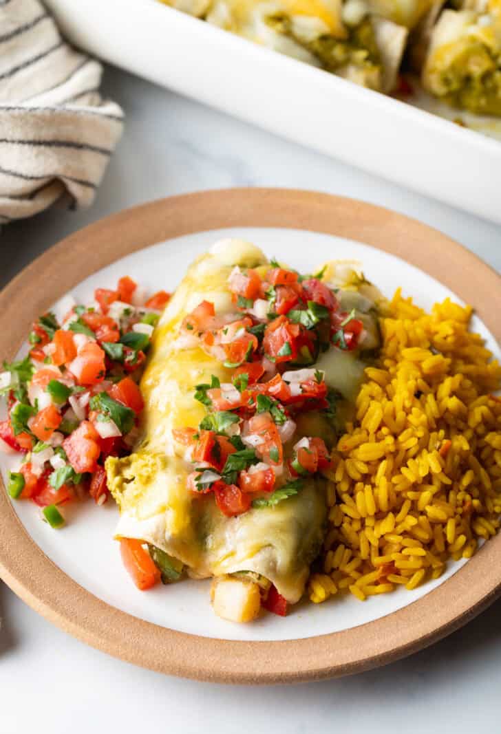 White plate with serving of pico de gallo, two chicken enchiladas with green sauce and pico de gallo, and spanish rice.