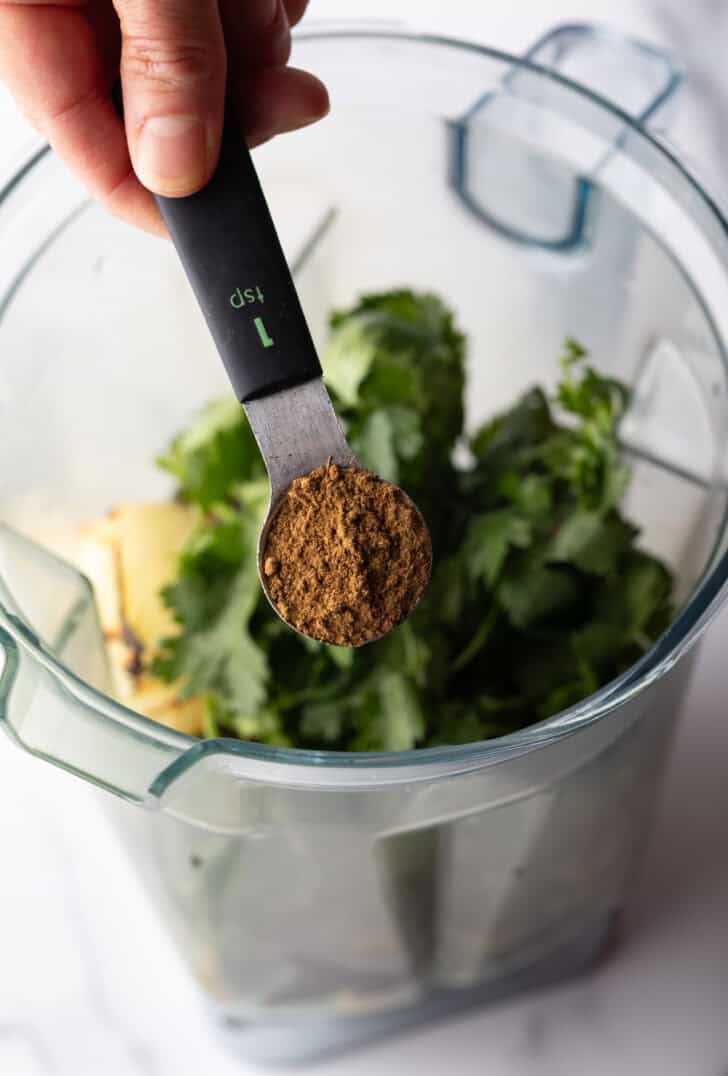 Hand with measuring spoon pouring spices into a clear blender pitcher with green herbs.