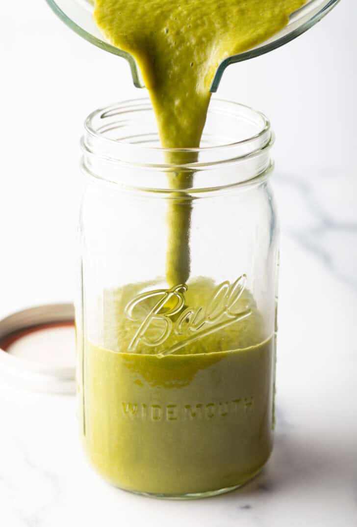 Pouring green sauce from a clear pitcher into a large wide mouth glass mason jar.