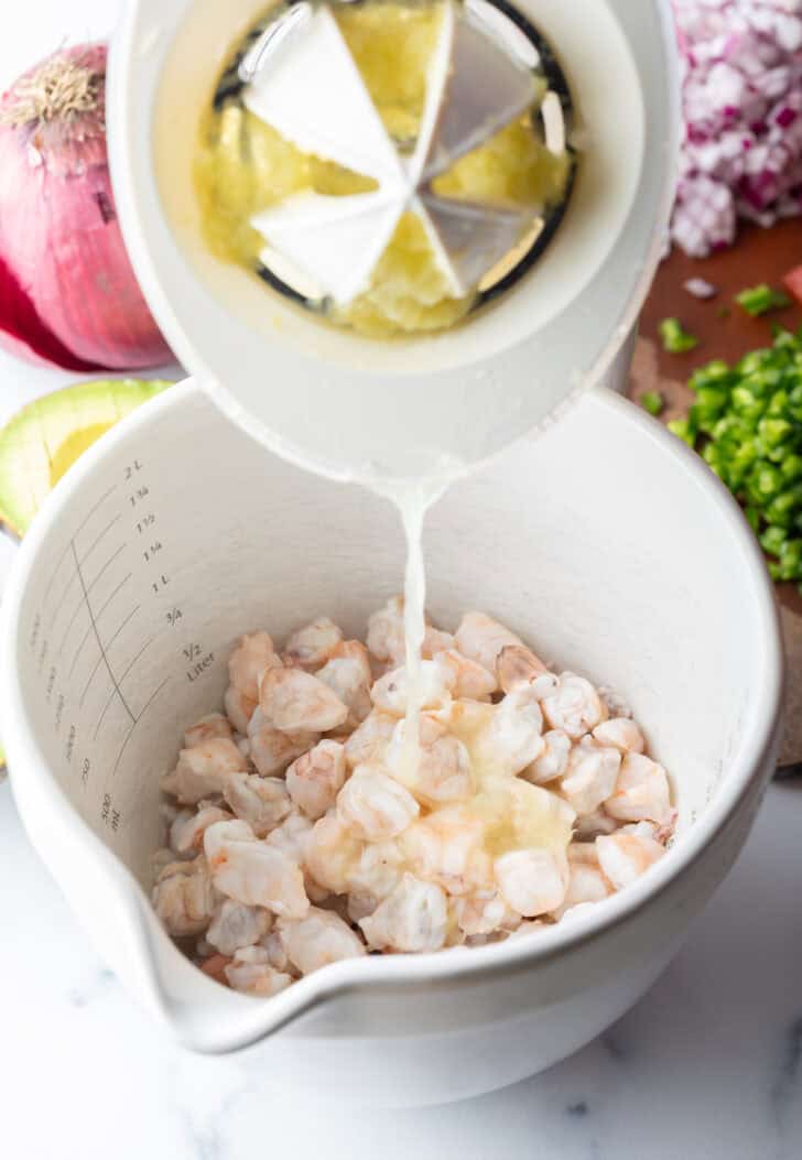 Pouring lime juice from a juicer into a white bowl of raw shrimp.