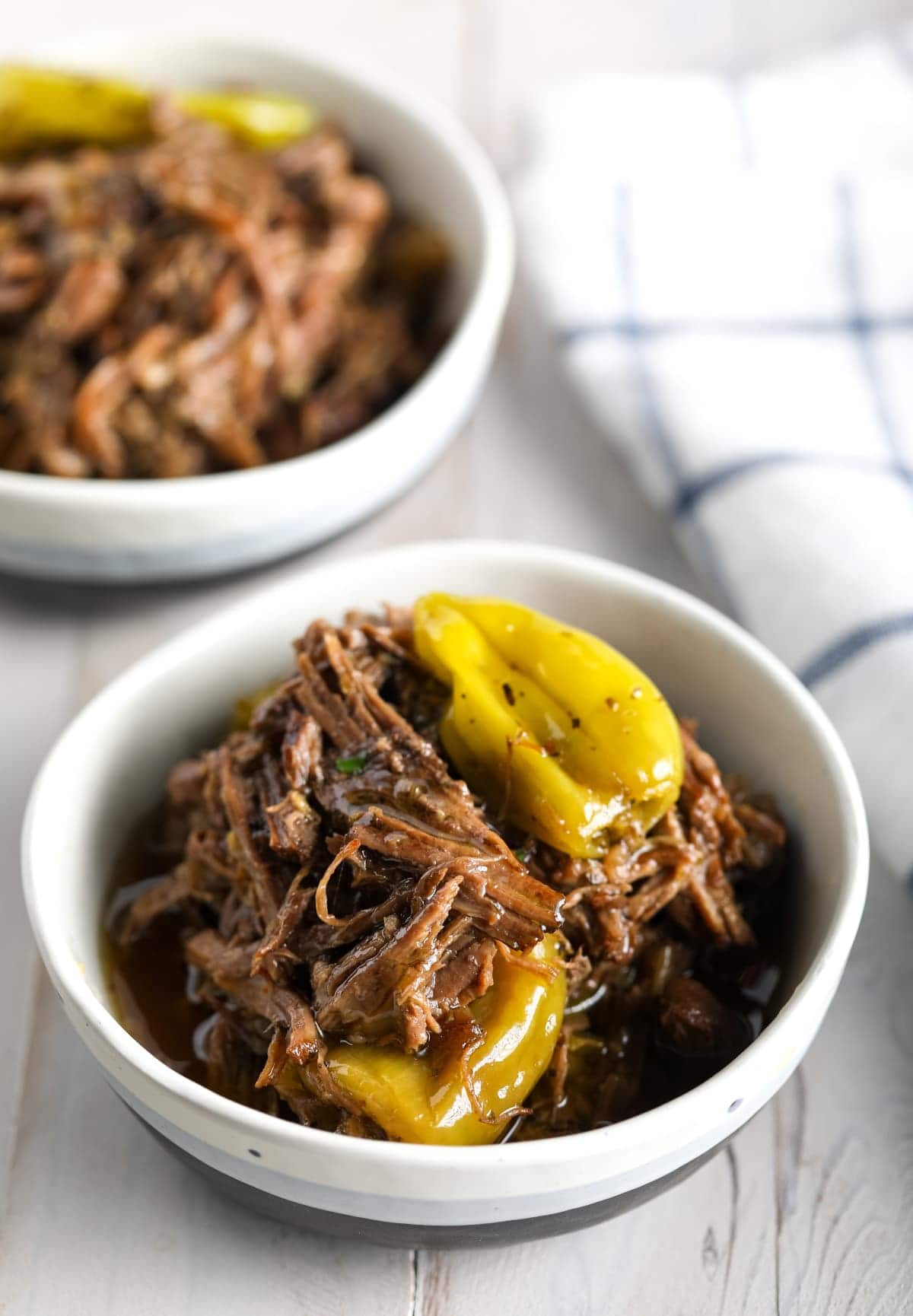 Two white bowls with Italian beef in them with pepperoncini peppers. 
