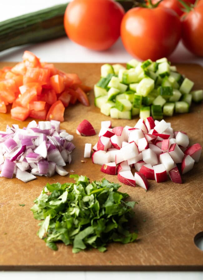Chopping board with recipe ingredients chopped and in piles: Cucumber, tomato, radish, red onion, and cilantro.