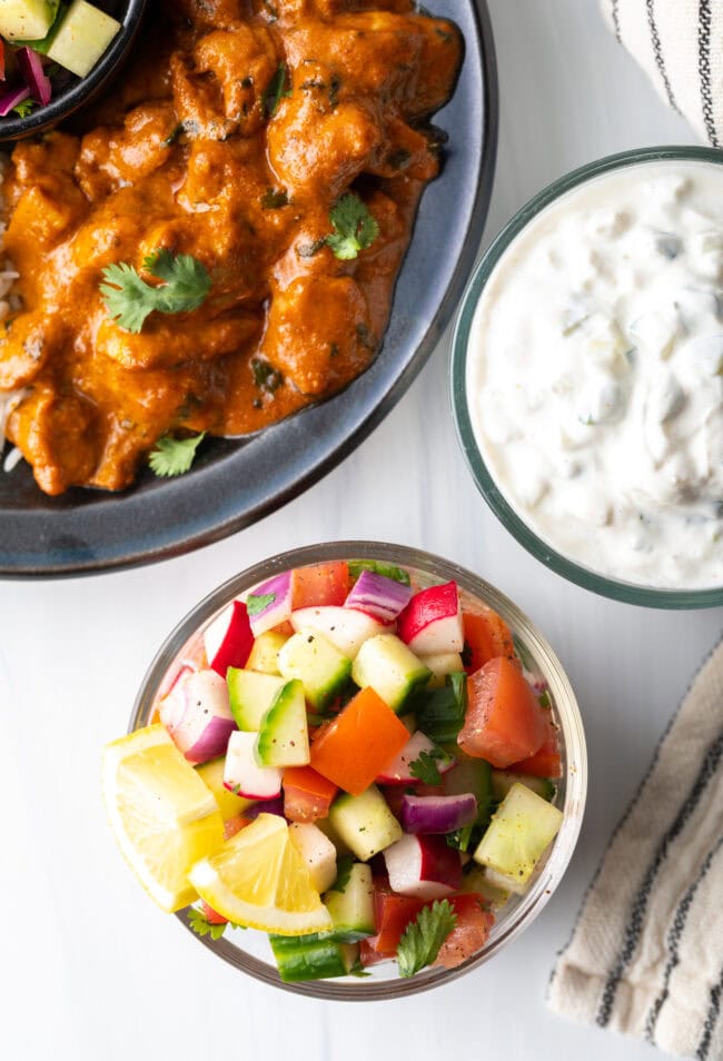 Top down view kachumber India salad in a glass bowl. There is also a glass bowl with raita and a dish with butter chicken.