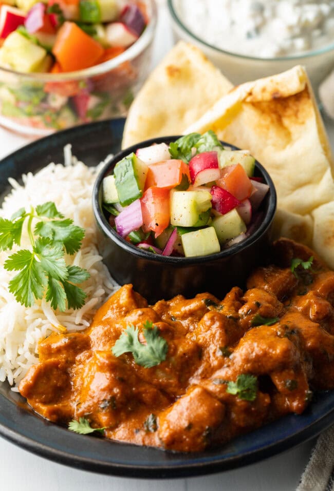 Serving of kachumber Indian salad in a black ramekin. The dish is on a larger black plate, surrounded by other Indian dishes including butter chicken, white rice, and naan.