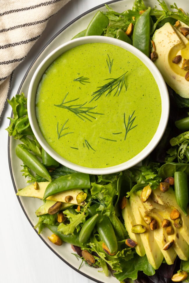 Top down view plate of salad with green, snap peas, avocado, and pistachios, and bowl of vegan homemade green goddess dressing on the side of the plate.