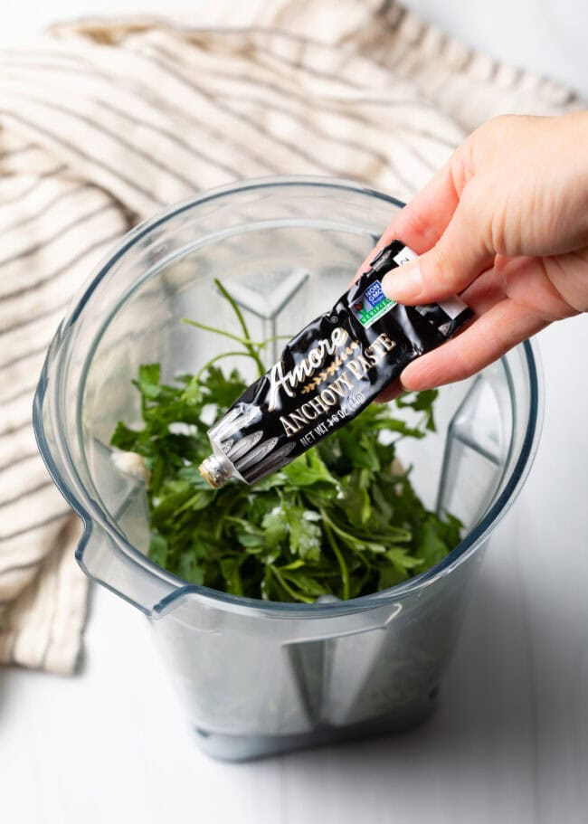 Hand squeezing a tube of anchovy paste into blender jar.